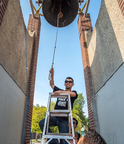 Chris is standing on a ladder underneath a large bell, ringing the bell with a smile.
