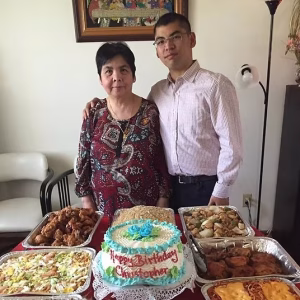 Chris stands with his mother in front of a beautiful spread of Filipino food and a cake that reads, Happy Birthday Christopher.